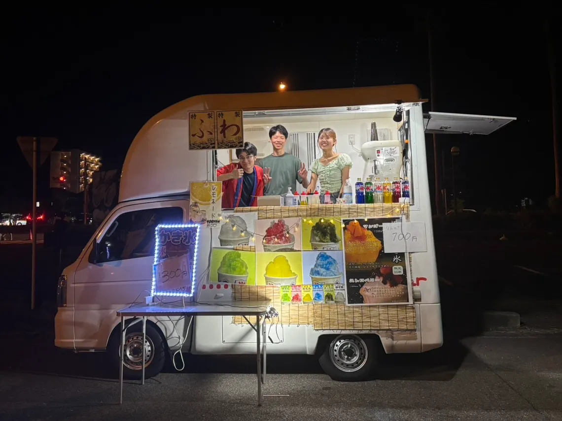 Our shaved-ice food truck lit up at night at the Kamegawa Summer Festival, with Pann and two teammates in the window